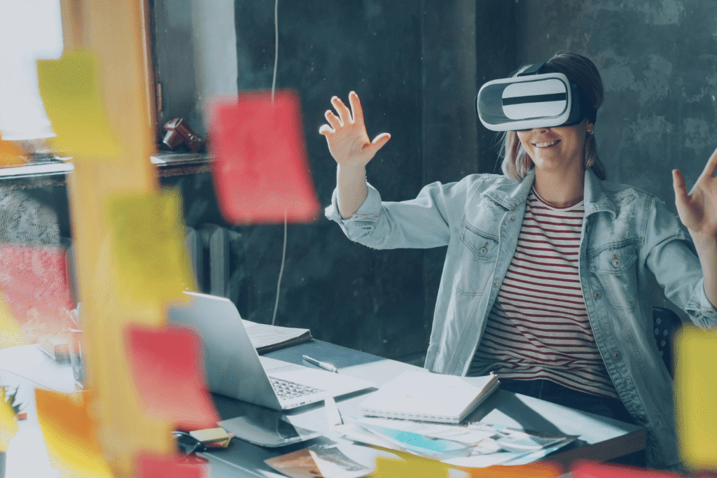 Modern workspace scene showing professional using AR/VR headset with raised arms interacting in immersive environment at desk with laptop and notes