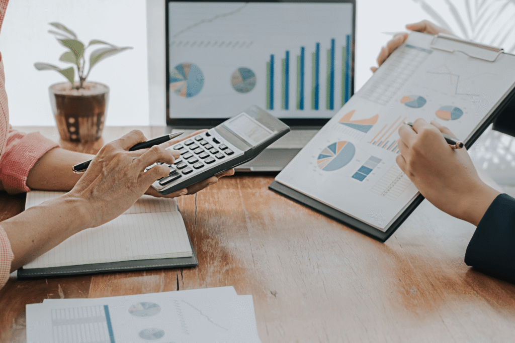 Office teamwork scene showing professionals using calculator, charts, and laptop dashboards to analyze financial data and business performance metrics