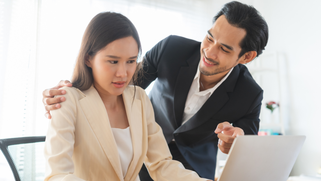 An office scene where a female employee appears visibly uncomfortable as a male colleague places his hand on her shoulder while leaning in and smiling. The woman looks distressed while working on her laptop, portraying an instance of unwanted behaviour in a professional environment.