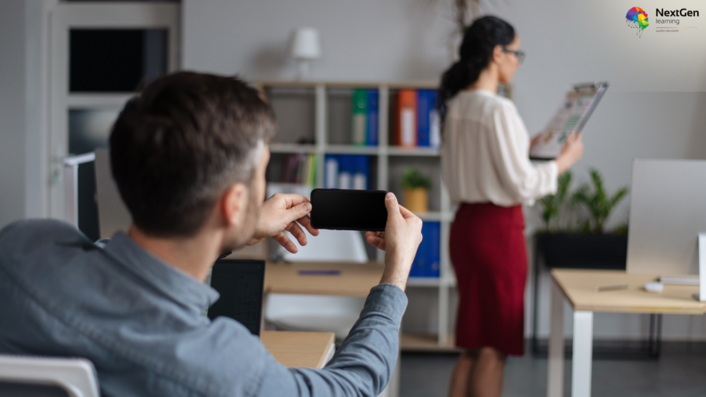 An office environment where a male employee discreetly takes a photograph on his smartphone of a female colleague standing at a distance, reviewing a clipboard. The image highlights an instance of inappropriate behaviour and invasion of privacy in the workplace. The NextGen Learning logo is visible in the top-right corner.