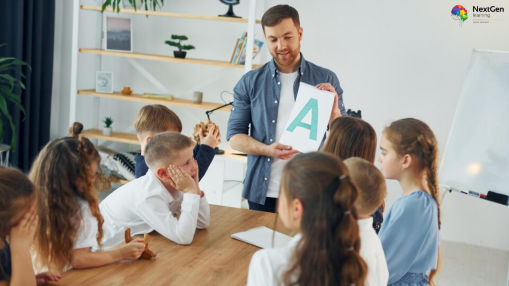 A male teacher showing a large flashcard with the letter 'A' to a group of attentive primary school children in a well-decorated classroom.