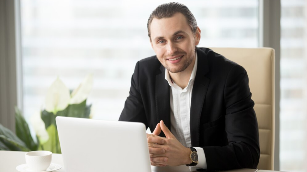 Confident investment banker in a suit smiling at his desk in a modern office setting, with a laptop and coffee cup.