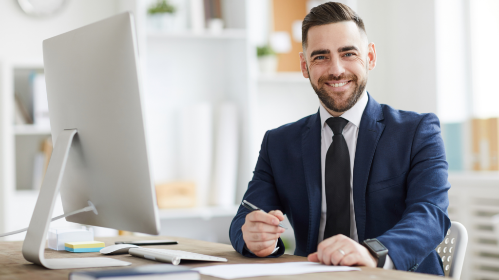 Smiling professional man in a navy suit seated at a desk in a modern office, holding a pen and facing a computer screen.