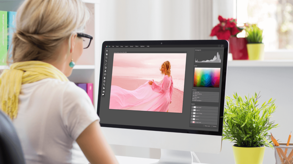 A woman wearing glasses and a yellow scarf works on photo editing software on a desktop computer, adjusting a vibrant image of a woman in a flowing pink dress on a beach.
